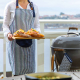 Femme portant le tablier de cuisine assorti à côté d’un barbecue avec une manique suspendue visible, sans personnalisation appar