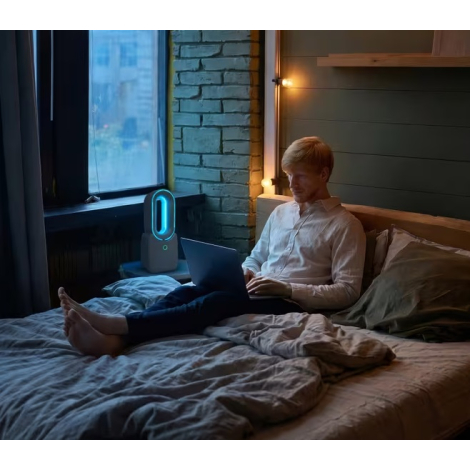 Homme utilisant un ordinateur portable sur un lit avec ventilateur sans pales allumé en arrière-plan, sans marquage visible.