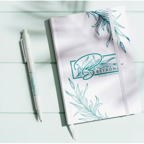 Carnet A5 présenté en situation d’utilisation avec marquage visible sur la couverture avant, posé à côté d’un stylo assorti.
