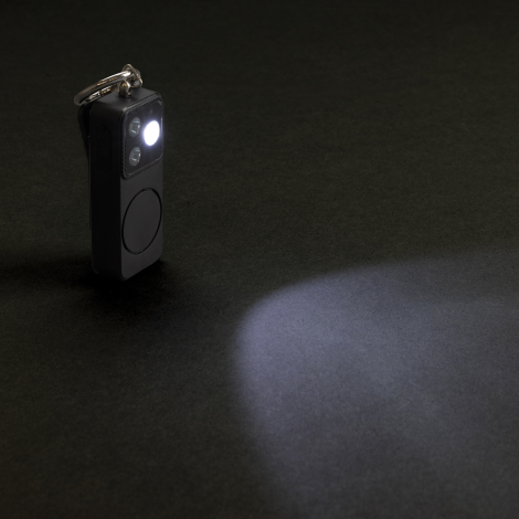 Porte-clés alarme posé sur une surface sombre avec éclairage LED activé, projetant un faisceau lumineux.