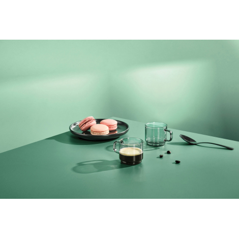 Mise en situation de deux tasses expresso sans marquage en verre fumé sur une table verte, dont une remplie de café. Une assiett