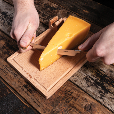 Personne utilisant le coupe-fromage pour découper un morceau de fromage posé sur la planche en bois, sans marquage apparent.