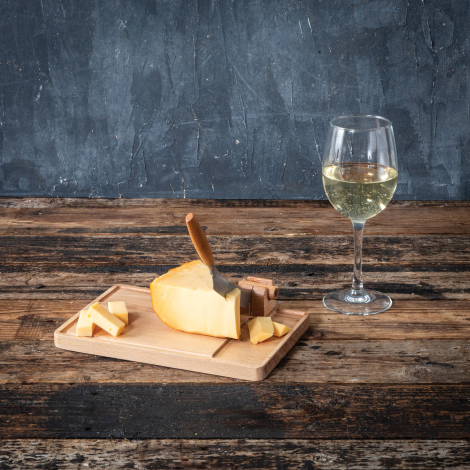 Coupe-fromage en situation d'utilisation avec un morceau de fromage sur la planche en bois et un verre de vin blanc à proximité,