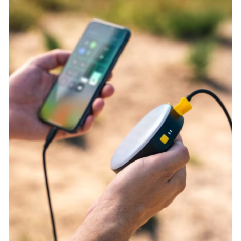 Lampe et batterie externe tenues en main et reliées à un smartphone en cours de charge dans un environnement extérieur.