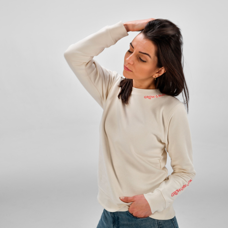 Une femme porte un sweat à col rond écru présenté de trois quarts de devant, avec un marquage visible au niveau du col et sur la