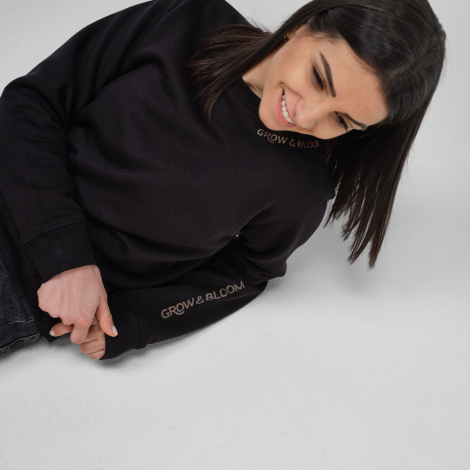 Une femme porte un sweat à col rond noir avec un marquage visible au niveau du col et sur la manche gauche.