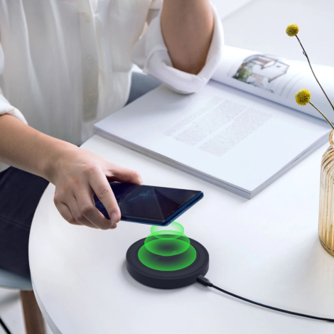 Chargeur rond noir posé sur une table blanche, relié à un câble, avec un smartphone tenu au-dessus et un effet graphique vert en
