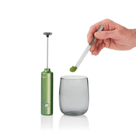 Mousseur électrique vert avec tige et fouet déployés, présenté à côté d’un verre transparent tandis qu’une main tient une cuillè