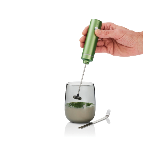 Mousseur électrique vert tenu à la main avec fouet plongé dans un verre contenant une boisson claire et de la poudre verte en su