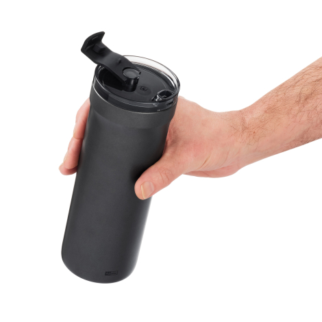 Mug isotherme noir tenu à une main et vu légèrement de dessus, avec couvercle transparent et système d’ouverture relevé.