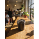 Tasse à café noire fermée posée sur une table en bois éclairée par le soleil, au premier plan d’un intérieur de café avec mobili