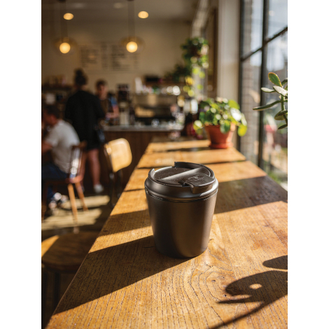 Tasse à café noire fermée posée sur une table en bois éclairée par le soleil, au premier plan d’un intérieur de café avec mobili