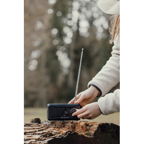Femme manipulant une radio multifonctions posée sur une souche en extérieur, avec antenne déployée, tandis qu'elle règle la comm