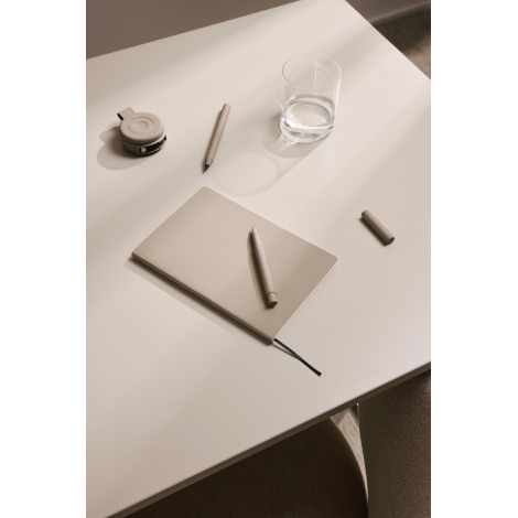 Carnet de notes beige fermé posé sur une table avec un stylo, un verre d’eau et de petits accessoires, son ruban marque-page noi