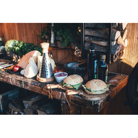 Râpe conique en métal avec poignée et support en bois présentée sur une table garnie de fromages, pains, condiments et accessoir