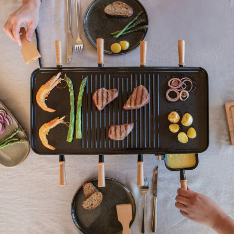 Appareil à raclette utilisé à table, avec viandes, crevettes, asperges, pommes de terre et oignons disposés sur la plaque de cui