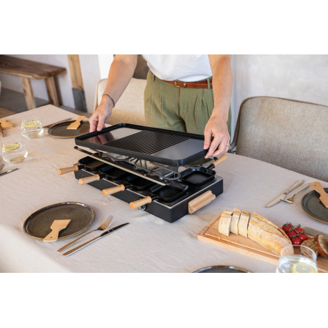 Appareil à raclette vu de trois quarts pendant son installation sur une table, montrant la plaque supérieure, les poêlons et les