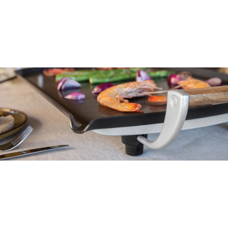 Gros plan sur le rebord de la plaque de cuisson et sur une poignée latérale en bois, avec aliments en cuisson sur la surface.