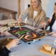 Plancha électrique utilisée à table par plusieurs personnes, avec viande, asperges, oignons et crevettes en cuisson sur la plaqu