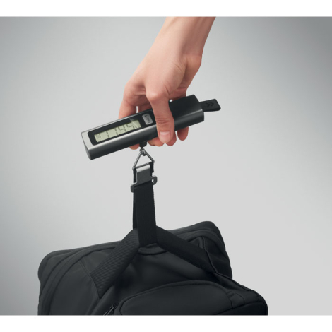 Utilisation du pèse-bagages pour soulever un sac noir par sa poignée, la sangle étant fixée au bagage tandis que le poids s’affi