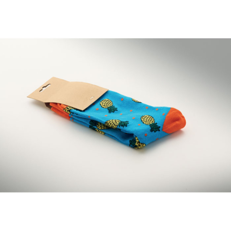 Paire de chaussettes pliée sous une étiquette en papier kraft, avec fond bleu turquoise, motifs d’ananas jaunes et verts et poin