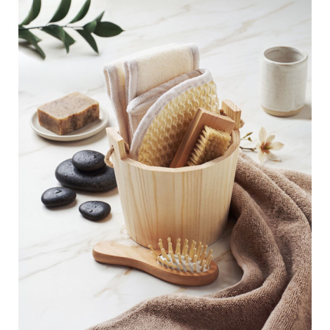 Le coffret de bain est mis en scène sur une surface claire avec ses accessoires disposés dans le baquet en bois, tandis qu’une p
