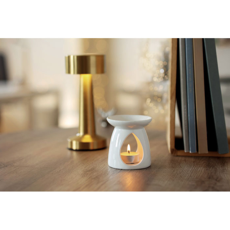 Le brûleur de parfum est mis en scène sur un meuble en bois, avec la bougie allumée visible dans son ouverture, à côté d’une lam