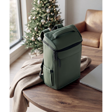 Sac à dos vert kaki posé sur une table dans un intérieur, à côté d’un ordinateur portable, avec sa poignée supérieure, ses poche