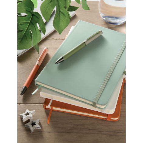 Plusieurs carnets A5 colorés sont empilés sur une table, avec un carnet vert au-dessus accompagné de deux stylos, tandis que des