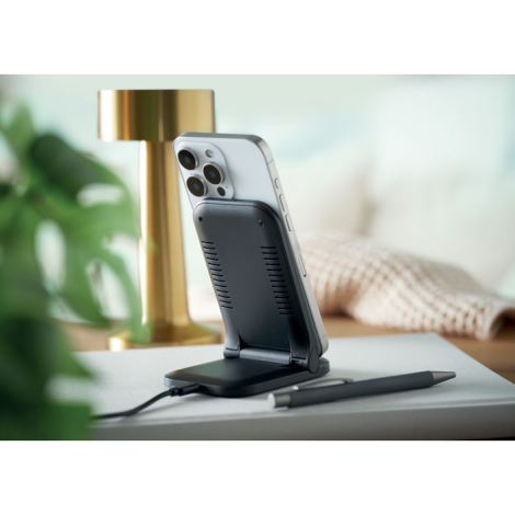 Posé sur un bureau, le chargeur noir maintient un smartphone verticalement sur son support incliné, à côté de deux stylos et d’u