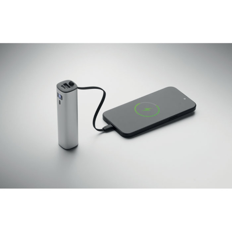 La batterie externe argentée est posée verticalement et reliée par son câble noir intégré à un smartphone placé à côté, dont l’é