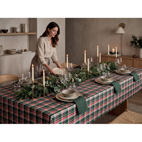 Une femme dresse une longue table recouverte de la nappe à carreaux verts, rouges et blancs, entourée de vaisselle, de verres, d