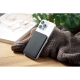 Smartphone blanc posé sur un tissu gris avec batterie externe noire fixée au dos, dans une mise en scène sur une table en bois a