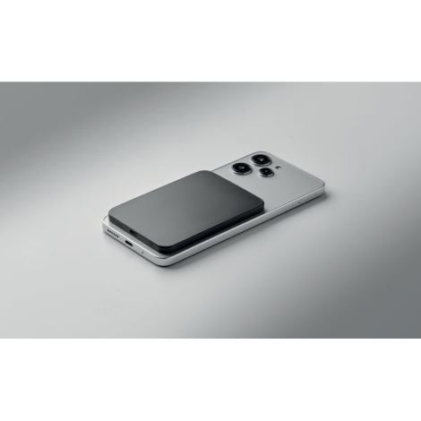 Smartphone blanc posé à plat avec batterie externe noire fixée au dos, présenté en perspective sur une surface grise.