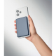 Smartphone blanc tenu en main avec batterie externe bleu foncé rectangulaire fixée au dos de l’appareil.