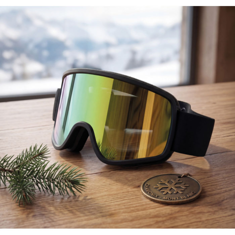 Masque de ski noir à écran miroir posé sur une table en bois devant un paysage de montagne enneigé, accompagné d’une branche de 