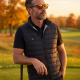 Homme portant un gilet chauffant noir matelassé sans manches sur un polo noir, avec col montant ouvert et bouton de commande sur Homme portant un gilet chauffant noir matelassé sans manches sur un polo noir, avec col montant ouvert et bouton de commande sur