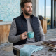 Homme assis à une table en bois, portant un gilet chauffant noir sans manches ouvert sur un pull gris, avec matelassage horizont Homme assis à une table en bois, portant un gilet chauffant noir sans manches ouvert sur un pull gris, avec matelassage horizont