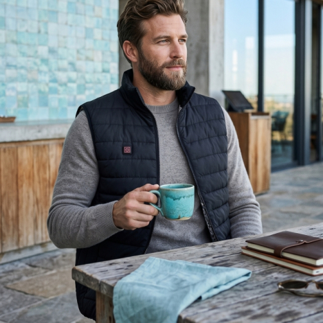 Homme assis à une table en bois, portant un gilet chauffant noir sans manches ouvert sur un pull gris, avec matelassage horizont