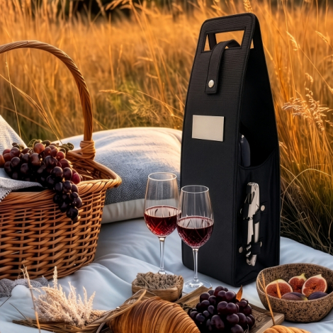 Sac porte-bouteille noir posé sur une nappe de pique-nique dans un champ doré, avec plaque métallique vierge et tire-bouchon lat