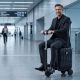 Un homme en costume sombre est assis sur une valise-trottinette noire dans un terminal d'aéroport, les mains sur le guidon en T 