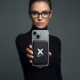 Une femme portant des lunettes noires et un haut à col montant présente un smartphone tenu verticalement, équipé au dos d'une ba