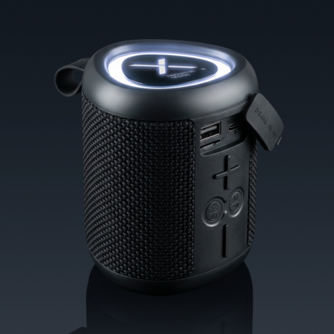 Enceinte Bluetooth noire photographiée de trois quarts sur fond sombre, avec grille textile tressée, boutons de commande, ports 