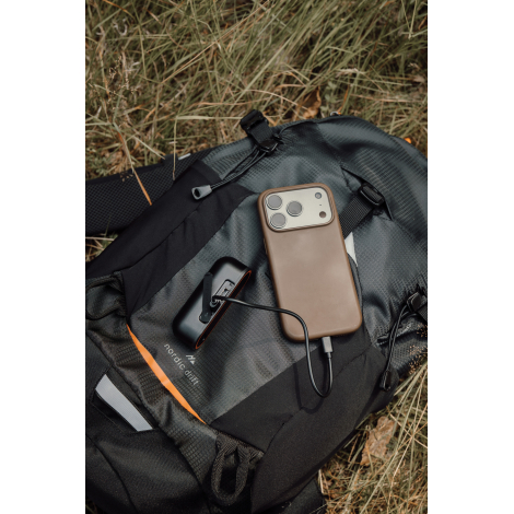 Un chauffe-main noir relié par un câble à un smartphone marron est posé sur un sac à dos gris et noir, dans l'herbe.