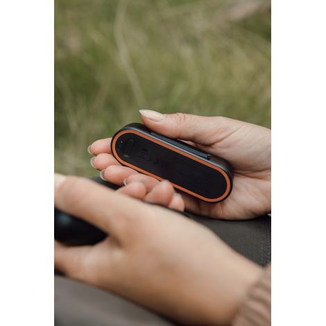 Un chauffe-main noir est tenu dans une main, montrant sa base entourée d'orange, son bouton ovale et trois voyants lumineux bleu