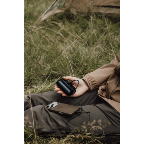 Une personne assise dans l'herbe tient un chauffe-main noir relié par un câble à un smartphone posé sur ses jambes.