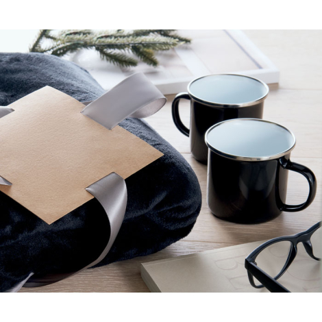 Mise en scène d'une couverture noire pliée avec ruban gris et étiquette en carton, accompagnée de deux mugs émaillés noirs sur u