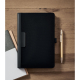 Conférencier A5 noir fermé, posé sur une table en bois à côté d'un stylo beige et de plusieurs carnets partiellement visibles. Conférencier A5 noir fermé, posé sur une table en bois à côté d'un stylo beige et de plusieurs carnets partiellement visibles.