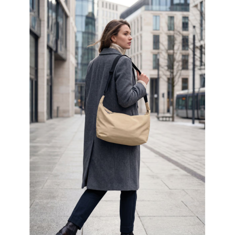 Une femme portant un manteau gris marche dans une rue urbaine avec un sac beige souple porté à l'épaule, muni d'une large bandou