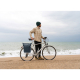 Un homme portant un bonnet vert se tient à côté d'un vélo sur une plage, face à la mer. Un sac à dos bleu foncé est fixé à l'arr Un homme portant un bonnet vert se tient à côté d'un vélo sur une plage, face à la mer. Un sac à dos bleu foncé est fixé à l'arr